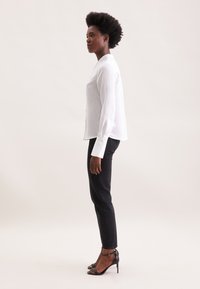 Camicia bianca a maniche lunghe con bottoni e righe verticali, abbinata a jeans skinny neri e tacchi a stiletto neri, vista di lato.