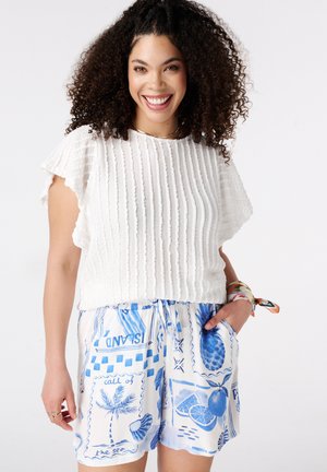 Lachende vrouw met krullend haar die een witte, getextureerde korte mouw top en witte shorts met blauwe tropische prints draagt, hand in de zak.