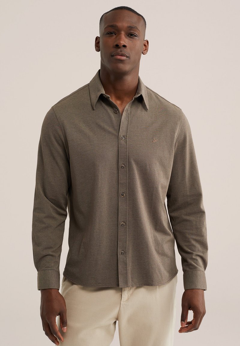 Homme portant une chemise marron à manches longues boutonnée et un pantalon beige, debout devant un fond clair uni.