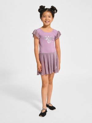 Młoda dziewczyna z fryzurą w podwójne koczki, ubrana w fioletową sukienkę do tańca z srebrnym motywem pszczoły i czarne baletki, uśmiechająca się do kamery.