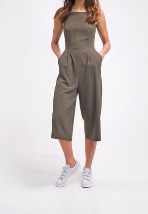 Olijfgroene jumpsuit met een vierkante halslijn, geplooide voorkant en zijzakken; gecombineerd met witte sneakers met drie banden.