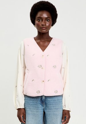 Donna con capelli corti e ricci che indossa un gilet rosa con bottoni floreali sopra una camicetta a maniche lunghe color crema e jeans blu, su uno sfondo semplice.