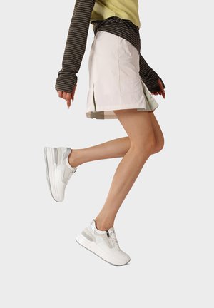 Persona che indossa scarpe da ginnastica bianche, gonna beige con spacco laterale e maglie a maniche lunghe marroni e gialle a strati, posando con una gamba sollevata.