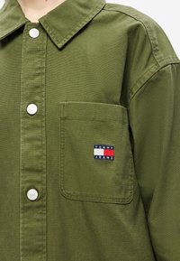 Πουκάμισο σε ελιά π�ράσινο με κουμπιά στο μπροστινό μέρος, μεγάλο γιακά και τσέπη στο αριστερό στήθος με μικρό λογότυπο "Tommy Jeans" σε κόκκινο, άσπρο και μπλε.