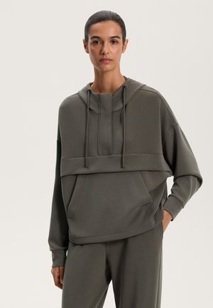 Sweatshirt à capuche surdimensionné en vert olive doux, présentant un design superposé, des cordons de serrage réglables et une poche avant. Texture lisse.