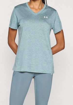 Person trägt ein hellblaues Under Armour Kurzarm-V-Neck-Shirt und passende Leggings, steht mit den Händen hinter dem Rücken.