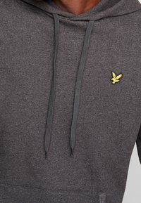 Ciemnoszara bluza z kapturem, wykonana z teksturowanej tkaniny, z przednią kieszenią, troczkami i małym żółtym logotypem orła na klatce piersiowej.