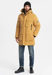 Gele waterdichte parka met vier voorzakken, capuchon en grijze reflecterende accenten; gecombineerd met blauwe jeans en zwarte veterschoenen.