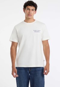 Młody mężczyzna z krótkimi kręconymi włosami ubrany w białą koszulkę Guess Jeans i niebieskie dżinsy, stoi na tle jasnego, jednolitego tła.
