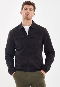 Zwarte lichte jas met een ritssluiting aan de voorkant, twee borstzakken en elastische manchetten. De stof is glad met een matte afwerking.
