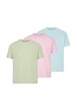 Femme aux cheveux bouclés portant un t-shirt vert clair oversize et un short en jean bleu clair retroussé, debout devant un fond uni.