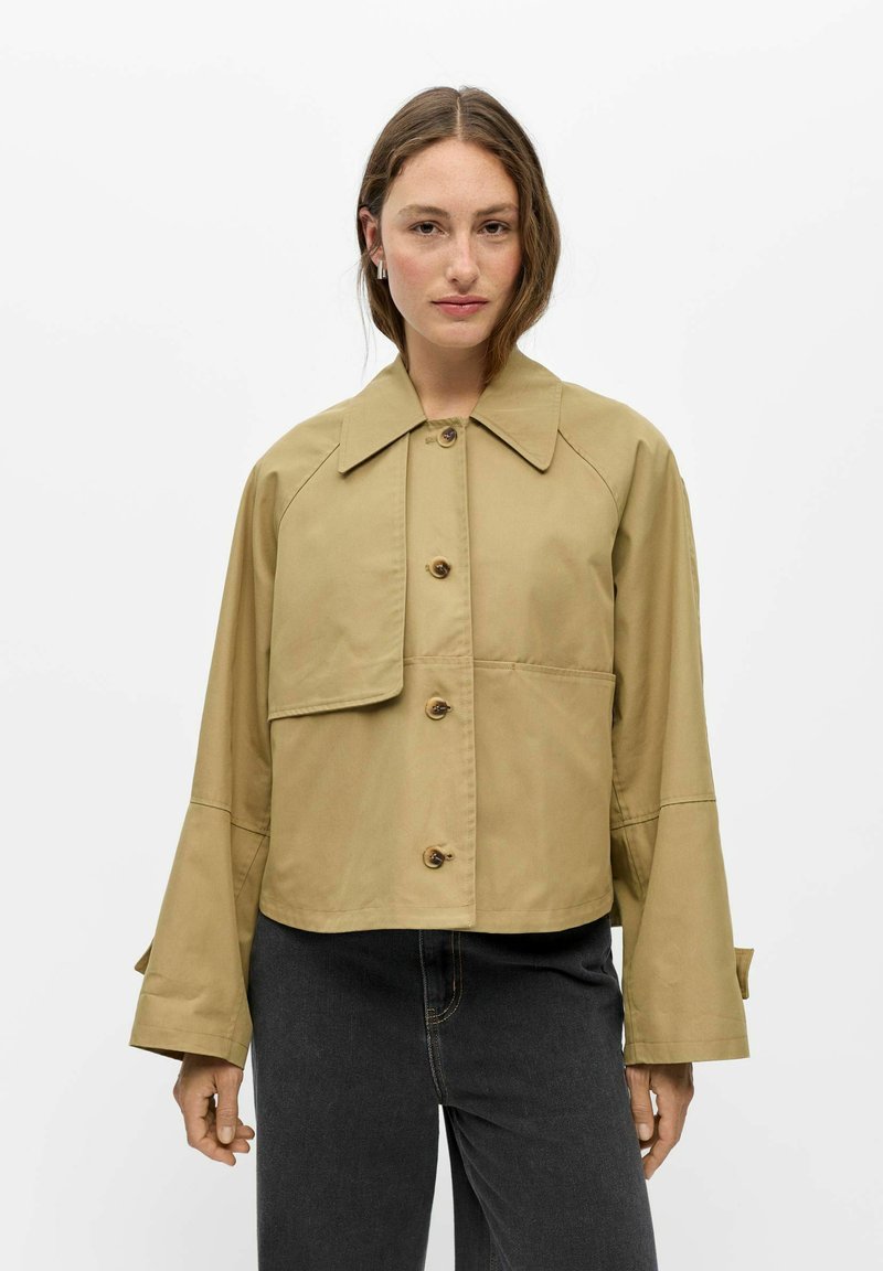 Femme portant une veste beige boutonnée avec de larges poignets et un grand col, associée à un jean sombre taille haute, debout sur un fond blanc.
