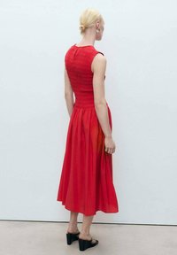Robe rouge avec un corsage plissé, une jupe fluide et un ourlet au genou. Le mannequin porte des mules noires à talons et a une coiffure lisse et stylisée.