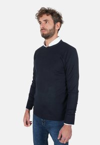 Maglione blu navy lavorato a maglia con scollo rotondo e polsini a coste, indossato sopra una camicia bianca con colletto. La texture appare liscia.