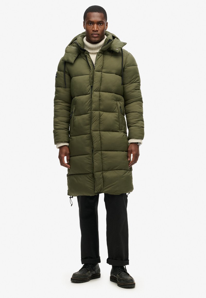 Oliven grøn pufferjakke med hætte, der har et quiltet design, frontlynlås og to sidelommer. Båret over en cremefarvet turtleneck og sorte bukser.