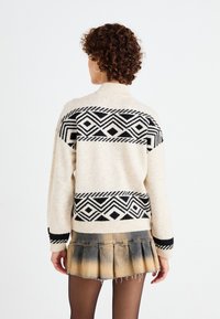 Femme aux cheveux bouclés portant un pull crème avec des motifs géométriques noirs et une jupe mini plissée, décolorée, noire et beige, de dos.