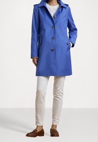 Abrigo azul tipo gabardina con botones al frente, cuello con muesca y bolsillos laterales, combinado con pantalones beige claro y mocasines marrones. Tejido suave y ligero.