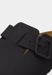 Μαύρο σανδάλι με φαρδιά ρυθμιζόμενη λουρίδα και ματ τετράγωνη αγκράφα, που δείχνει μέρος της ταμπά εσωτερικής επένδυσης και υφή στην σόλα.