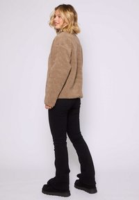 Kobieta z blond włosami stoi bokiem, ubrana w brązową polarową kurtkę, czarne dopasowane spodnie i czarne buty na platformie, na tle jednolitego tła.