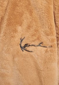 Kurtka z beżowego futra ekologicznego o gładkiej fakturze i czarnym haftowanym logo. Projekt jest elegancki z miękkim, puszystym wykończeniem.