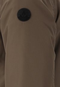 Brun vattentät jacka med en slät yta, med en cirkulär svart patch med texten "WHISTLER" på övre vänstra sidan.