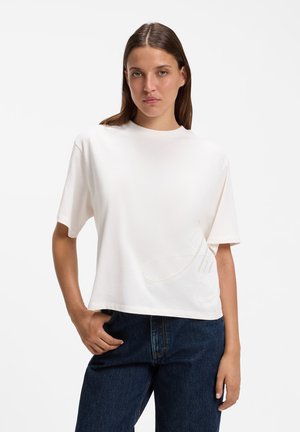 Ung kvinde med lige brunt hår iført en løs hvid kortærmet skjorte og mørkeblå jeans står med den ene hånd i lommen.