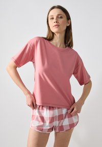 Tricou scurt din material moale, de culoare roz, asociat cu pantaloni scurți cu carouri roz și albe. Tricoul are o croială lejeră și un decolteu rotund.