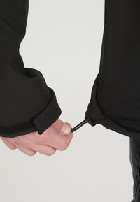 Black jacket with a textured finish; close-up of a hand pulling a drawstring at the cuff; features a Velcro flap and elastic binding.