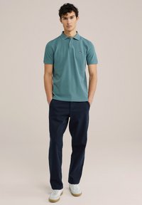 Polo de color verde azulado con dos botones y logo en el pecho, combinado con pantalones navy y zapatillas blancas con suela de goma.