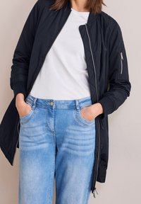 Kobieta w czarnej kurtce bomberce zapinanej na zamek, nosząca białą koszulę oraz jasnoniebieskie dżinsy, z rękami w kieszeniach spodni, stojąca na tle jednolitej ściany.