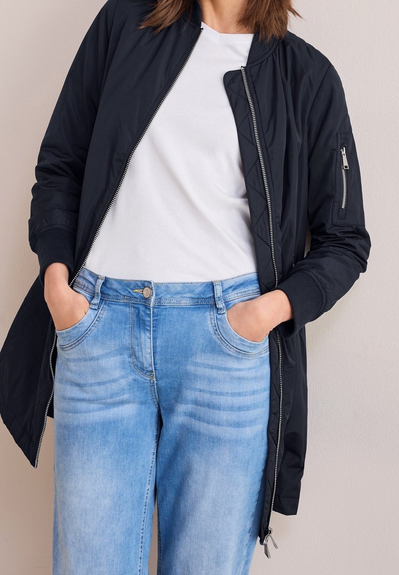 Kobieta w czarnej kurtce bomberce zapinanej na zamek, nosząca białą koszulę oraz jasnoniebieskie dżinsy, z rękami w kieszeniach spodni, stojąca na tle jednolitej ściany.