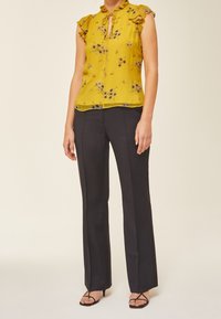 Femme portant un chemisier sans manches jaune à motifs floraux, un pantalon noir sur mesure et des sandales à talons noires à lanières, debout devant un fond uni.