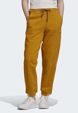 Personne portant un pantalon de jogging jaune moutarde avec des cordons noirs et blancs et des baskets blanches, mains dans les poches.