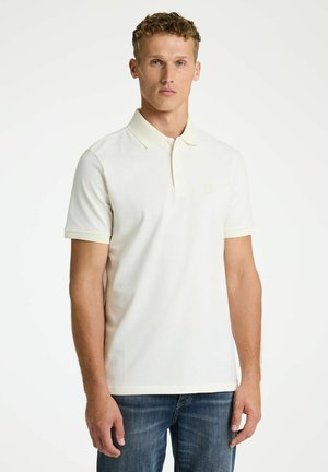 Fiatal férfi rövid, göndör hajú, egyszínű fehér rövid ujjú pólót és kék farmert visel, fehér háttér előtt áll.