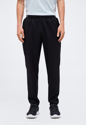 Mann trägt schwarze elastische Bundhose und schwarze Sportschuhe, steht vor einem einfarbigen hellen Hintergrund.