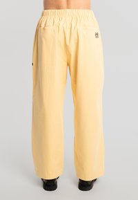 Persona vistiendo pantalones sueltos de tiro alto en color amarillo pálido con cintura elástica y un pequeño bordado floral negro en el bolsillo trasero.