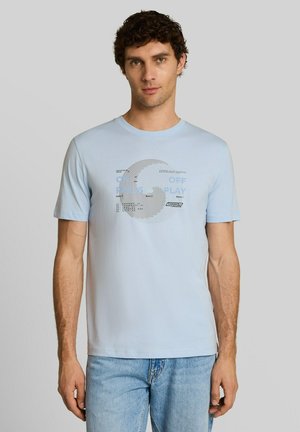 Mann mit lockigem Haar trägt hellblaues T-Shirt mit rundem grafischem Design und hellblaue Jeans und steht vor einem einfarbigen Hintergrund.