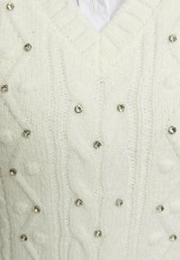 Maglione bianco lavorato a maglia con scollo a V, caratterizzato da un motivo a treccia e decorato con pietre decorative rotonde in tonalità argento.
