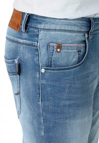 Light blue denim jeans featuring a tapered fit, multiple pockets, and a brown leather tag on the waistband. Subtle fading and stitching details.