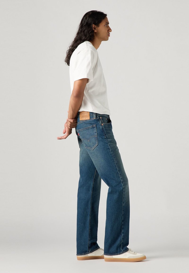 Blå denimjeans med rak bendragning, en läderlapp på baksidan av midjan, i kombination med en enkel vit T-shirt och vita sneakers.