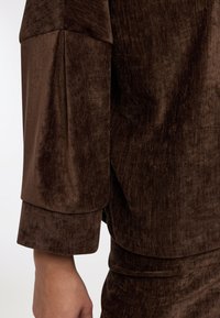 Tessuto marrone con texture e una morbida lucentezza, caratterizzato da una manica larga e rilassata e un design a strati sull'orlo. Aspetto liscio e vellutato.