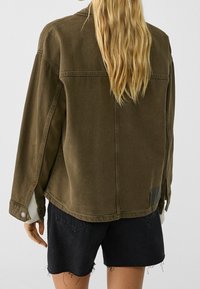 Blondyn w oversize'owej oliwkowej kurtce i czarnych szortach dżinsowych z postrzępionymi brzegami, zwrócony plecami do kamery.