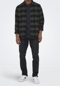 Chemise en flanelle à carreaux noirs et verts superposée sur un t-shirt noir, associée à un jean noir et des baskets blanches avec des accents verts.