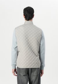 Grå vadderad väst med hög krage, med sidoknappar och en lätt design. Bärs över en ljusblå långärmad skjorta.