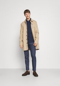 Beige lång jacka över en marinblå polo, kombinerad med blå denimbyxor och bruna mockaskor. Har detaljer med knappar och fickor med dragkedja.