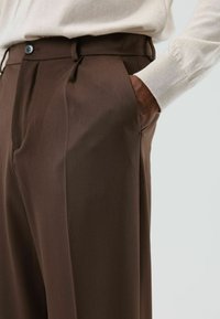 Bruine op maat gemaakte broek met een gladde textuur, plooien aan de voorkant, zijzakken en een knoopsluiting; gecombineerd met een lichte trui.