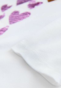 Tela blanca con costuras texturizadas. Se observan patrones de corazones morados que aportan un toque de color. Primer plano del borde de la manga y la costura de la prenda.