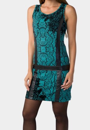 Frau trägt ein ärmelloses türkisfarbenes Kleid mit schwarzen floralen und geometrischen Mustern, kombiniert mit transparenten schwarzen Strumpfhosen, steht vor einem weißen Hintergrund.