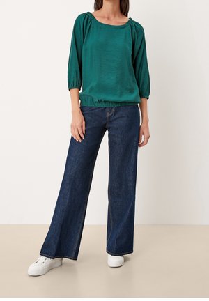 Kvinde iført mørkegrøn bluse med 3/4-ærmer, vidtbenede mørkeblå jeans og hvide sneakers, stående foran en ensfarvet baggrund.