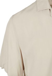 Chemise beige à manches courtes avec un col pointu, couture d'épaule visible et texture de tissu lisse.
